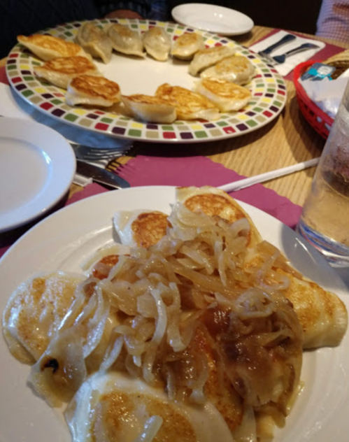 Two plates of pierogi, one topped with sautéed onions, served with a glass of water in a restaurant setting.