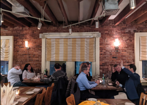 A cozy restaurant interior with brick walls, wooden beams, and diners engaged in conversation at candlelit tables.