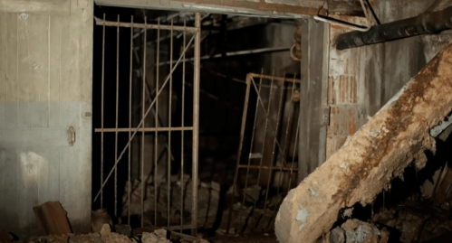 A dark, abandoned space with a rusty gate and debris scattered on the ground, suggesting decay and neglect.