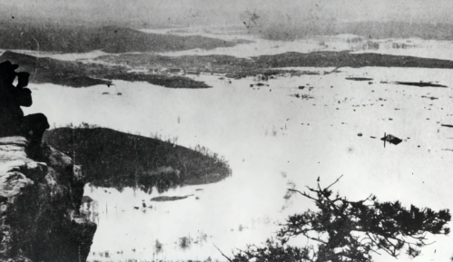 A black-and-white photo of a person sitting on a cliff, overlooking a foggy landscape with water and distant hills.