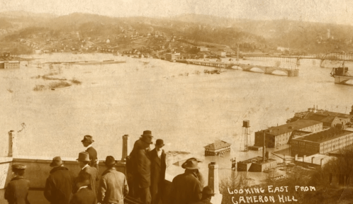 Historic photo showing a flooded landscape from Cameron Hill, with people observing the water and distant buildings.