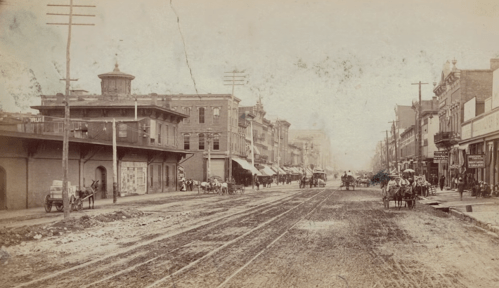 Historic street scene with dirt road, horse-drawn carriages, and buildings lining both sides, capturing a bygone era.