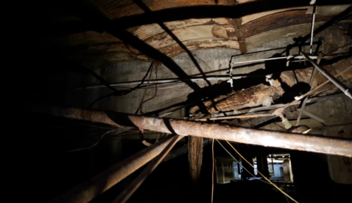 Dimly lit view of a dilapidated ceiling with exposed pipes and wooden beams, surrounded by tangled wires.