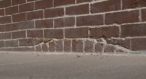 Close-up of a brick wall with visible wear and cracks at the base, showing a concrete floor beneath.