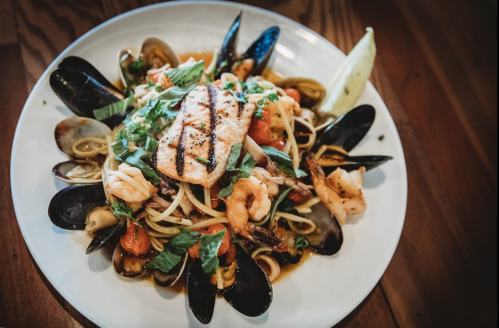 A plate of seafood pasta with shrimp, mussels, and grilled salmon, garnished with herbs and lemon.