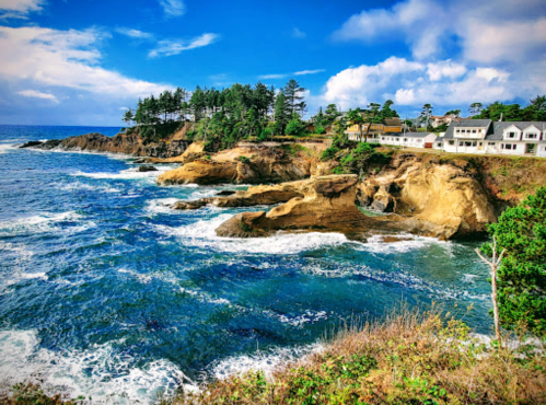 Coastal view with rocky cliffs, waves crashing, and houses nestled among trees under a blue sky with scattered clouds.