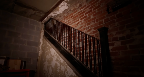 A dimly lit staircase with a black railing, leading up to a brick wall and peeling paint.