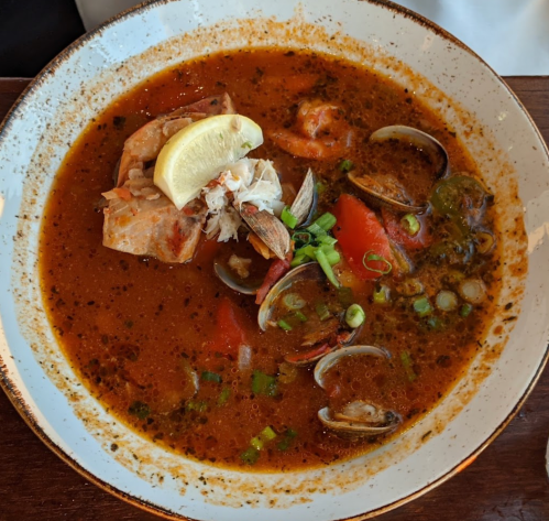 A bowl of seafood soup with clams, crab, and a lemon wedge, garnished with green onions and tomatoes.