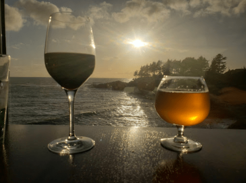 Two glasses of wine sit on a table overlooking a sunset by the ocean, with trees in the background.