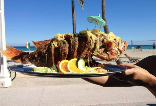 A large grilled fish garnished with onions, cucumber, and an umbrella, served on a platter at a beach.