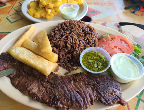 A plate of grilled steak with rice, fried plantains, tomato slices, and two dipping sauces on a colorful table.