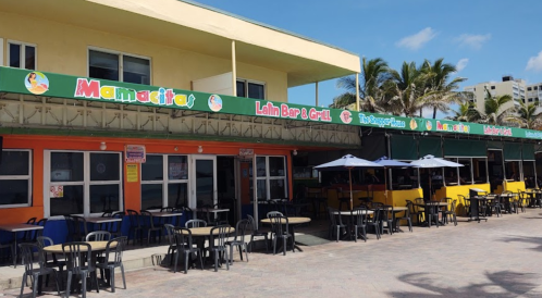 Colorful beachfront restaurants with outdoor seating and umbrellas under a clear blue sky.