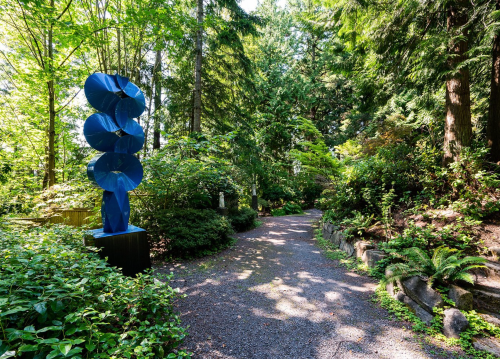 A winding path through a lush green garden, featuring a tall blue sculpture amidst trees and foliage.