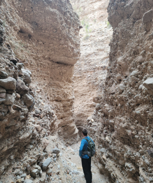 A person stands in a narrow canyon surrounded by steep, rocky walls. The landscape is dry and rugged.