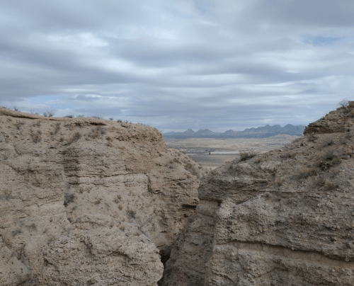A rugged canyon landscape with rocky cliffs and distant mountains under a cloudy sky.