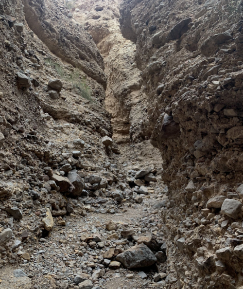 A narrow canyon with steep rocky walls and a dry, rocky floor, surrounded by loose stones and gravel.