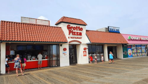 Grotto Pizza restaurant on a boardwalk, with people walking by and colorful shops nearby.