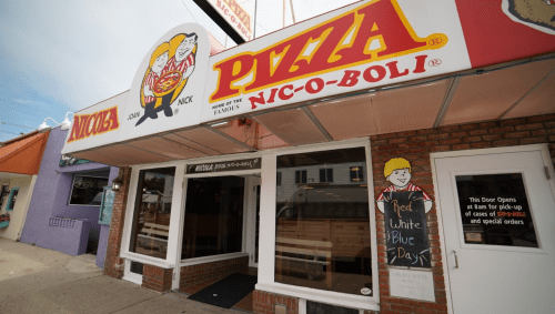 Exterior of a pizza shop with a colorful sign featuring a cartoon chef and the name "Nic-O-Boli."