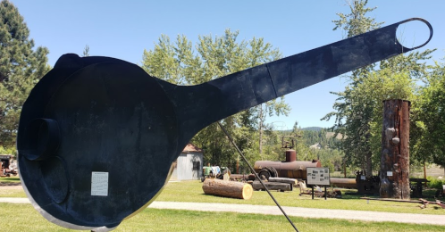 A large, black metal spoon sculpture stands in a grassy area, surrounded by trees and industrial artifacts.