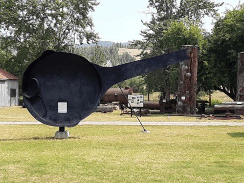 A large, black metal spoon sculpture stands in a grassy area, surrounded by trees and historical artifacts.