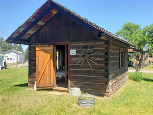 A rustic wooden cabin with a sloped roof, open door, and a sun decoration on the wall, surrounded by green grass.