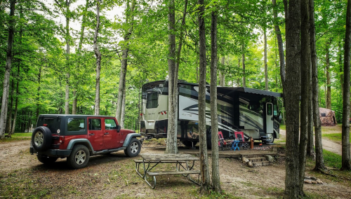 A red Jeep parked next to a large RV in a wooded campsite, surrounded by trees and a picnic table nearby.