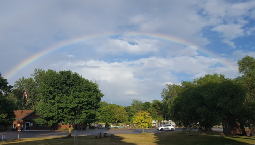 A vibrant rainbow arcs over a green landscape with trees and a parking area under a partly cloudy sky.