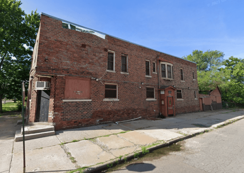 A weathered brick building with boarded windows and a red door, set on a quiet street surrounded by trees.