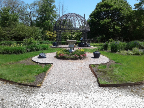 A serene garden featuring a decorative gazebo, a fountain, and lush greenery surrounded by gravel paths.