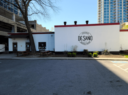 Exterior of DeSano Pizzeria, featuring a white building with a logo, picnic tables, and nearby greenery.