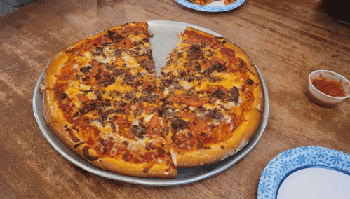 A pizza with pepperoni and sausage, partially sliced, on a metal tray with plates and dipping sauce in the background.