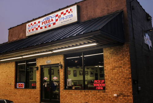 Exterior of The Pizza Plaza restaurant at dusk, featuring a lit "OPEN" sign and a red and white checkered awning.