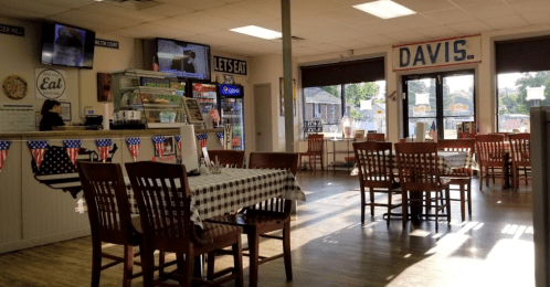 A cozy diner with checkered tables, a counter displaying snacks, and patriotic decorations. TVs are visible in the background.