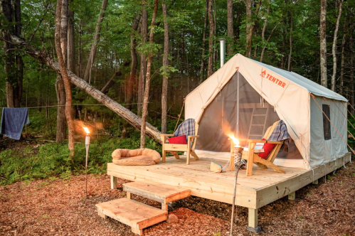 A cozy tent on a wooden deck in a forest, with chairs, a fire pit, and torches, surrounded by greenery.