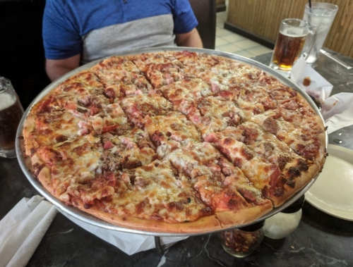 A large, round pizza topped with cheese, meats, and vegetables, served on a metal tray in a restaurant setting.