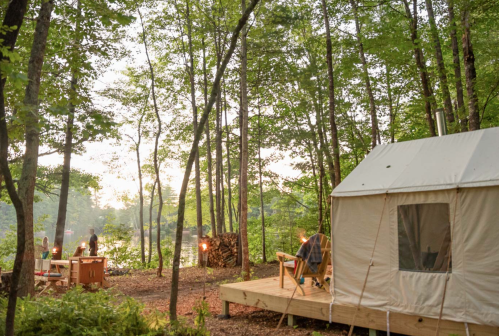 A cozy tent in a wooded area near a calm lake, with chairs and a fire pit set up for relaxation.