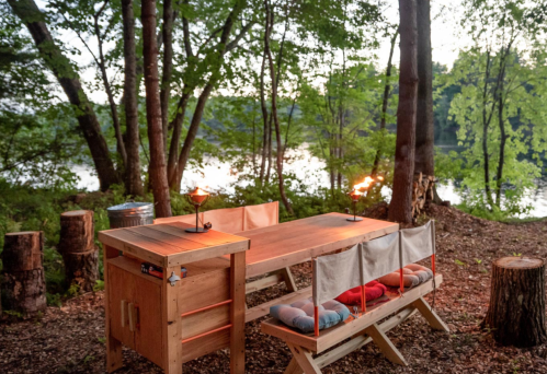 A wooden picnic table with cushions sits by a serene lake, surrounded by trees at dusk.