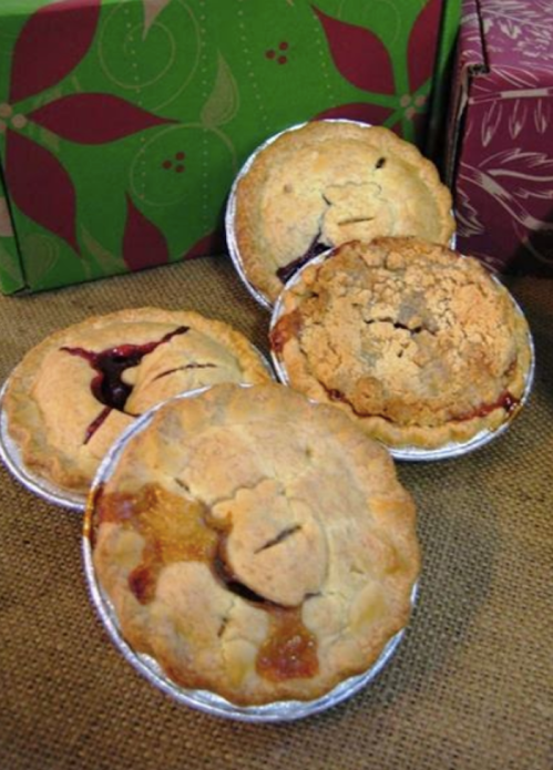 Four small fruit pies with golden crusts, placed on a burlap surface, surrounded by festive gift boxes.