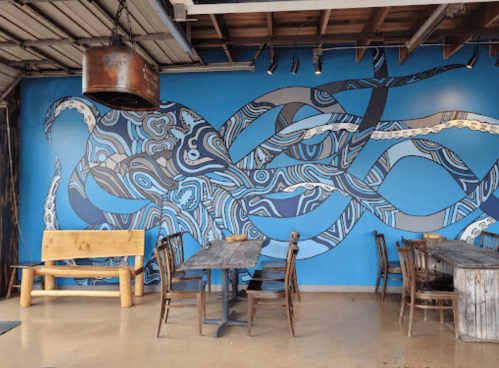 A vibrant blue mural of an octopus adorns a restaurant wall, with wooden tables and chairs in the foreground.