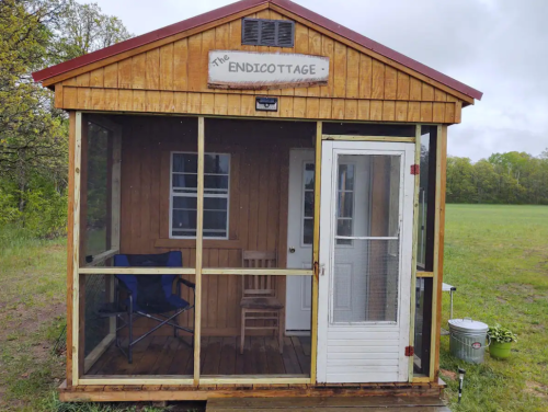 A small wooden cabin with a screened porch, featuring a sign that reads "The Endicottage." Surrounded by greenery.