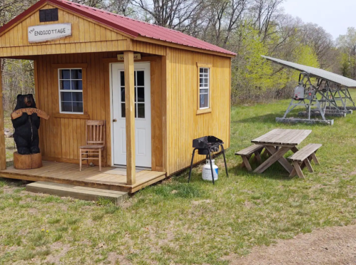 A wooden cabin with a red roof, a bear sculpture, picnic tables, and a solar panel nearby in a grassy area.