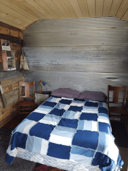 Cozy bedroom with a patchwork blue blanket on a bed, two wooden chairs, and a corrugated metal wall.