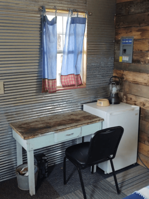 A small room with a wooden desk, chair, fridge, and a window with blue curtains. Walls are made of corrugated metal and wood.