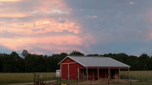 A red barn sits in a field under a colorful sunset sky with clouds, surrounded by trees.