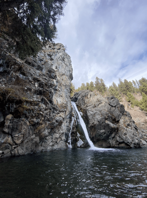 A serene waterfall cascading down rocky cliffs into a calm pool, surrounded by trees and a cloudy sky.