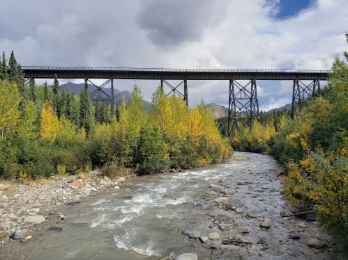 A tall railway trestle bridge spans a river, surrounded by vibrant autumn foliage and mountains under a cloudy sky.