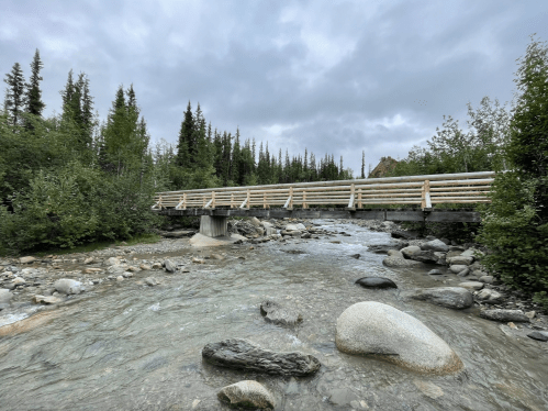 A wooden bridge spans a rocky river, surrounded by lush greenery and cloudy skies.