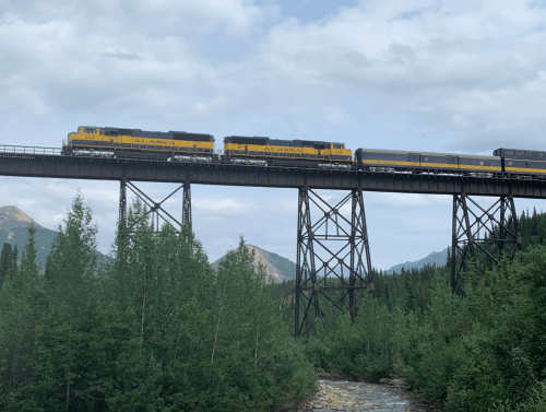 A yellow and black train crosses a tall trestle bridge surrounded by green trees and mountains under a cloudy sky.