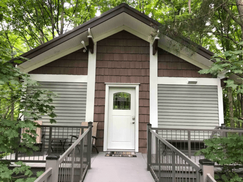 A cozy cabin with a brown and gray exterior, surrounded by lush green trees and featuring a welcoming front porch.