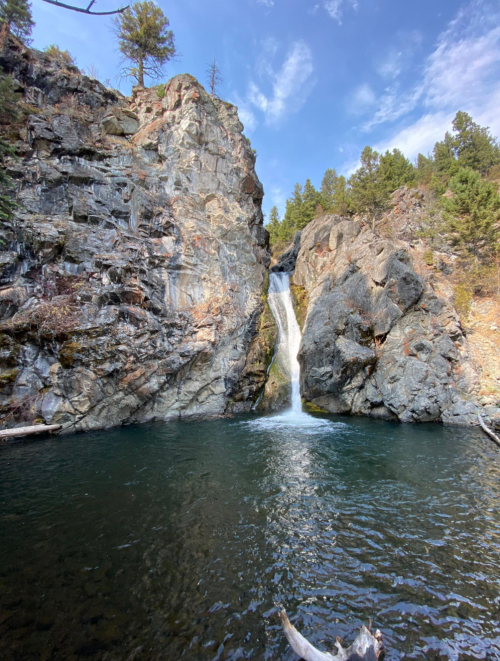 A serene waterfall cascades down rocky cliffs into a clear pool, surrounded by trees and blue sky.